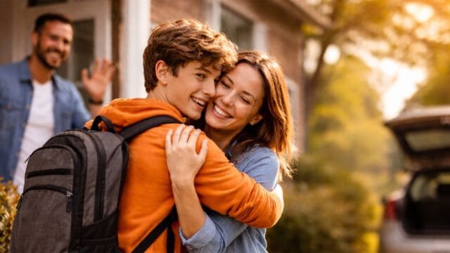 Teenager umarmt seine Mutter beim Abschied vor dem Wechsel zum anderen Elternteil im Nine-to-Nine-Lebensentwurf, Vater winkt im Hintergrund, warme herbstliche Umgebung, Patchworkfamilie in harmonischer Familiensituation