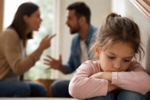 Im Bildhintergrund sieht man zwei Eltern streiten. Im Vordergrund sitzt ein Mädchen traurig auf der Treppe