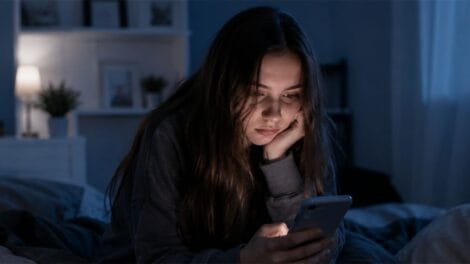 Ein Teenager sitzt auf dem Bett und scrollt mit dem Handy durch Soical Media. Ihr Gesichtsausdruck ist gelangweilt.