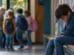 Ein traurig wirkender Junge sitzt allein auf einer Holzbank in einem Schulflur, während im Hintergrund eine Gruppe anderer Kinder miteinander zusammensteht.