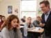 Ein Lehrer steht im Klassenzimmer und lacht über eine Schülerin, die sichtlich betroffen und traurig an ihrem Platz sitzt, während die Mitschüler im Hintergrund arbeiten.
