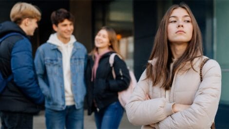Jugendliche vor einer Schule, während ein Mädchen selbstbewusst und abweisend abseits der Gruppe steht.