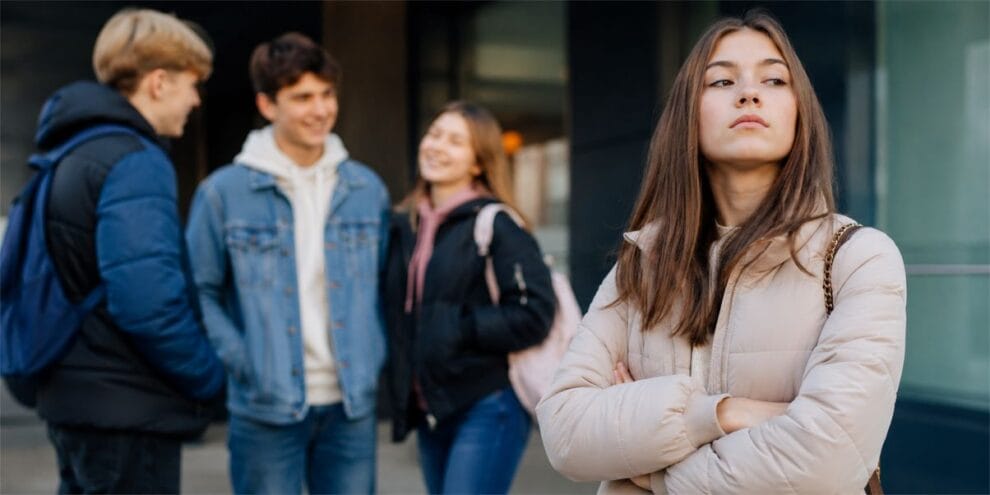 Jugendliche vor einer Schule, während ein Mädchen selbstbewusst und abweisend abseits der Gruppe steht.